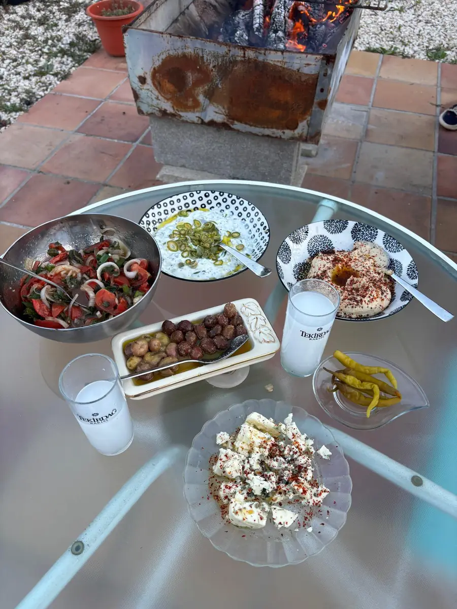 A table outside with salad, olives, yoghurt dip, hummus, feta and two glasses of rakı, by a fire